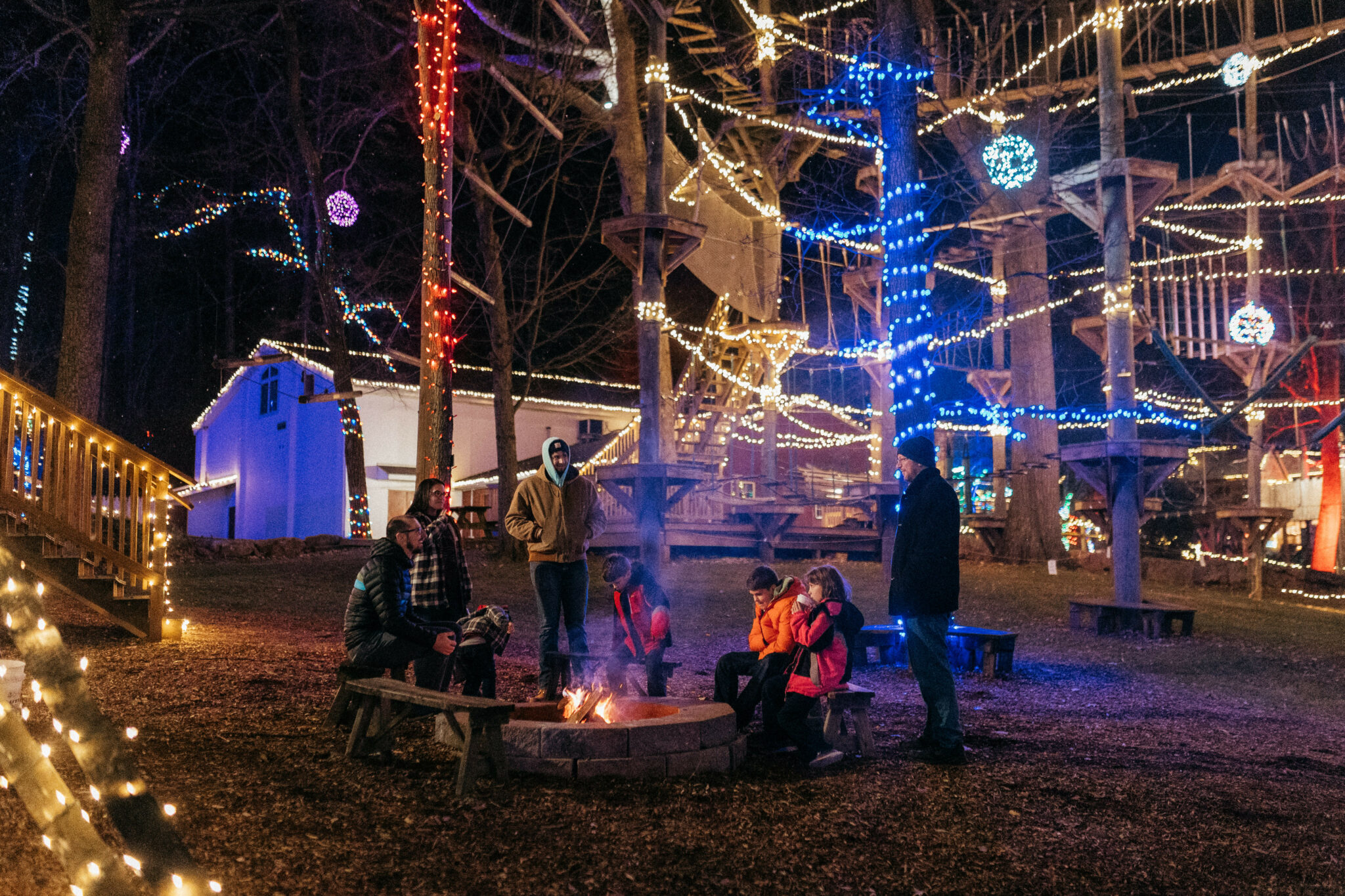 A Family Sitting At An Outdoor Fire While Lights Illuminateone Of Refreshing Mountain'S Christmas Zipline Experience